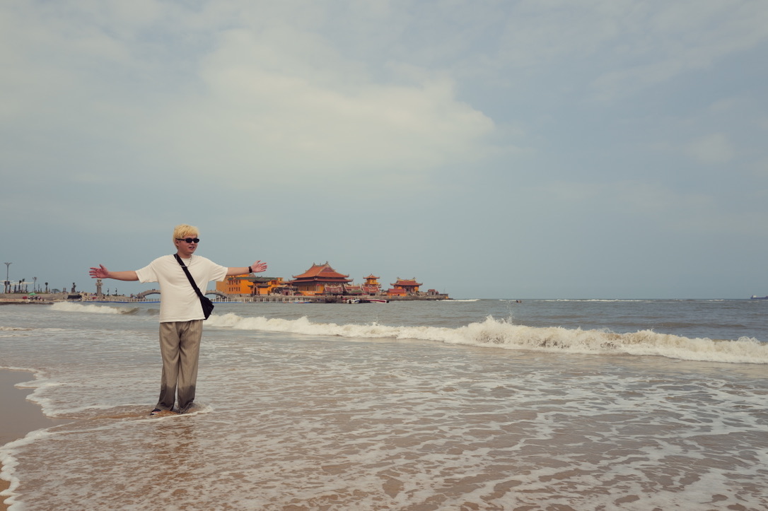 Xinyu Guan standing in the surf with a coastal temple in the distance