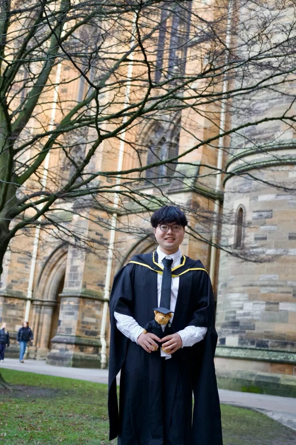 Xinyu Guan in graduation robes holding a teddy bear outside a University of Glasgow building